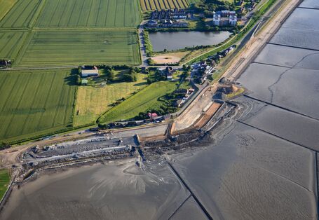 The picture shows an aerial view of a land protection dyke under construction. In the background, there are agricultural areas and houses as well as standing water visible. 
