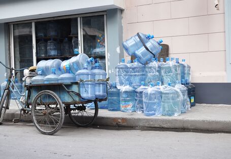 Altes Lastendreirad, welches auf der Straße abgestellt wurde und mit leeren Wasserkanistern vollbepackt ist; auf dem Fußgängerweg lagern weitere leere Plastikkanister; einige sind in Plastiktüten eingepackt, andere nicht; die Kanister kommen aus einem Lager, welches offen ist