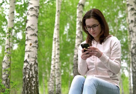Das Bild zeigt eine junge Frau, die in einem Birkenwwald sitzt. Sie blickt fokussiert auf ein Smartphone.