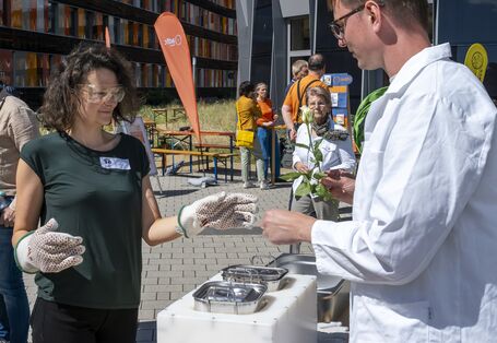 Die Umweltprobenbank hat es ermöglicht, mit einer Blattsammlung auf genetische Spurensuche gehen, Zu sehen sind ein Mann mit Laborkitel und Schutzbrille sowie eine Teilnehmerin mit Schutzbrille, der etwas aus einem Stickstoffbehälter gezeigt wird.. 