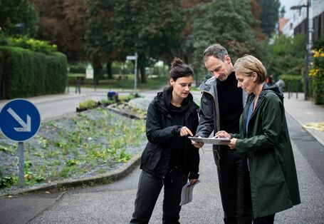 Der Wettbewerb Blauer Kompass stellt seine Bewerber vor, damit abgestimmt werden kann, welches Projekt gewinnt. Zu sehen sind drei Menschen der Berliner Regenwasseragentur, die auf der Straße stehen uns sich einen Plan angucken. 