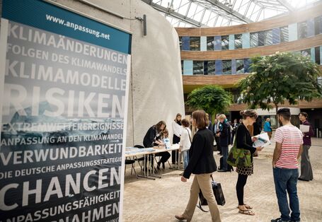 Blick ins Forum des Umweltbundesamt auf den Anmeldetisch. Personen befinden sich hinter und vor dem Anmeldebereich. Im Vordergrund steht ein Roll up beschriftet mit den Umweltrisiken und Chancen.