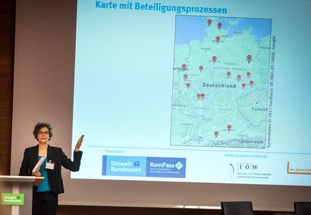 Vortragende steht am Rednerpult und zeigt auf Deutschlandkarte im Hintergrund. Die Karte zeigt an in Rot markiert, wo Beteiligungen bereits stattfinden.