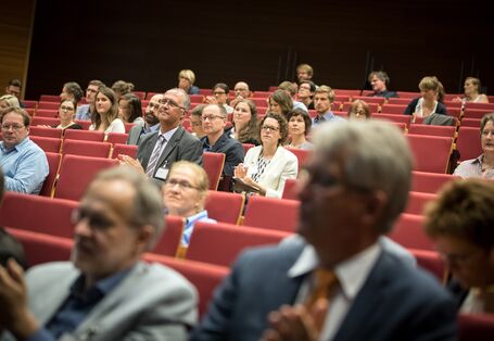 Blick von vorne in die Sitzreihen des Hörsaales. Veranstaltungsteilnehmerinnen und -nehmer sitzen zuhörend auf roten Reihenstühlen.