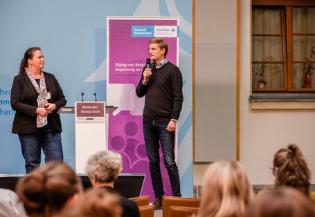 Zwei Teilnehmende stehen am Stehpult vor dem blauen Hintergrund, auf dem der Veranstaltungsort geschrieben steht sowie das lilafarbene Veranstaltungs-Rollup steht.