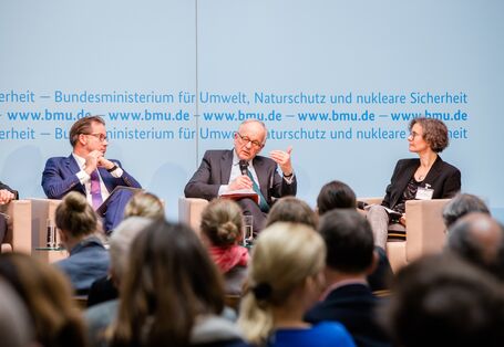 Podiumsdiskussion vor hellblauem Hintergrund, auf dem der Veranstaltungsort geschrieben steht.