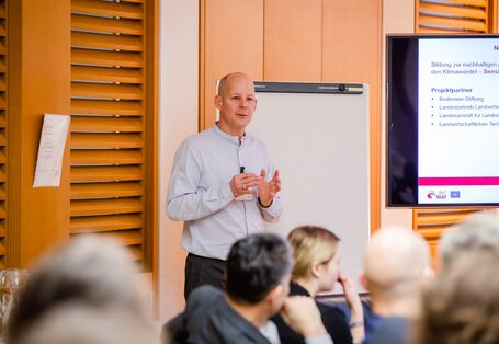 Teilnehmender steht vor einem leeren Flipchart und spricht gestikulierend zur Gruppe. Die Teilnehmenden sitzen in einem Raum, der mit holzfarbenen Elemente vertäfelt ist.