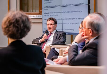 Podiumsdiskussion aus einer anderen Perspektive. Im Hintergrund steht eine Tafel, auf der die Podiumsbeteiligten verzeichnet sind.
