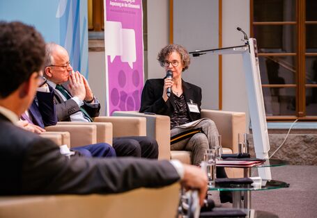 Podiumsdiskussion aus einer anderen Perspektive. Im Hintergrund steht ein Rollup in lila Farbtönen mit Veranstaltungstitel. Die Moderatorin spricht in das Mikrofon.