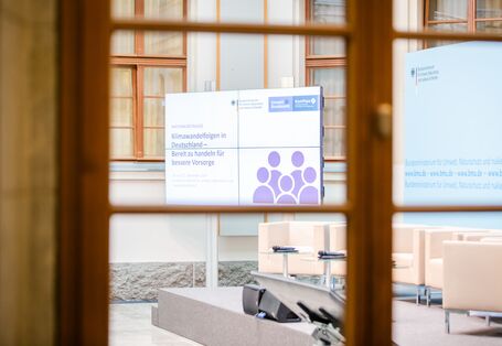 Blick durch das hölzerne Fensterkreuz eines Workshopraumes in den Innhof auf den Titelaufsteller sowie die Podiumsausstattung, wie Sessel, Tische, Mikrofone.