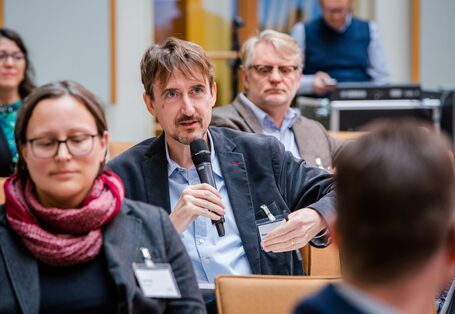 Ein Teilnehmer sitzt im Veranstaltungsraum mit den anderne Teilnehmenden und spricht ins Handmikrofon. 