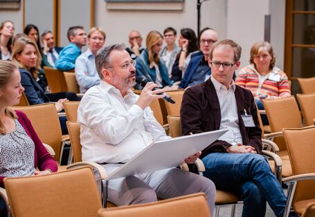 Teilnehmende sitzen im einem Veranstaltungsraum auf orangenen Stühlen in Reihen und blicken nachdenklich. Ein Teilnehmer spricht in ein Handmikro und hält ein Tableau in der Hand.