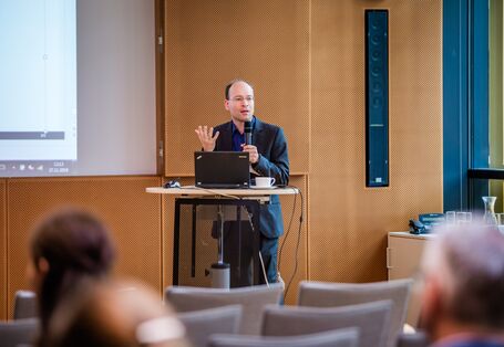 Ein Vortragender steht vor dem geöffnetem Laptop gestikulierend am Stehpult im holzfarben vertäfelten Veranstaltungsraum. Der Vortragende trägt einen dunkelfarbenen Anzug.