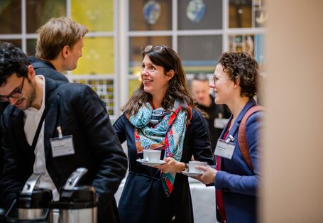 Teilnehmende stehen in der Pause mit Kaffetassen in den Händen beisammen und sprechen lachend miteinander.