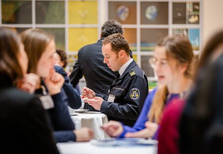Teilnehmende stehen in der Kaffeepause an Stehtischen zusammen und tauschen sich rege aus. Ein uniformierter Teilnehmender isst Kuchen.