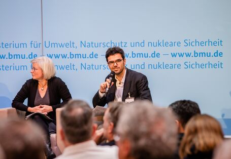 Podiumsdiskussion vor dem hellblauem Hintergrund, auf dem der Veranstaltungsort geschrieben steht. Ein Teilnehmender hält ein Handmikrofon.