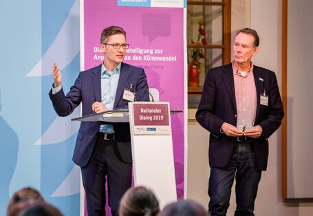 Teilnehmender trägt gestikulierend am Stehpult vor. Ein weiterer Teilnehmer steht dabei und hört zu. Im Hintergrund steht der Veranstaltungs-Rollup in lila Farbtönen mit Veranstaltungstitel und Logo.