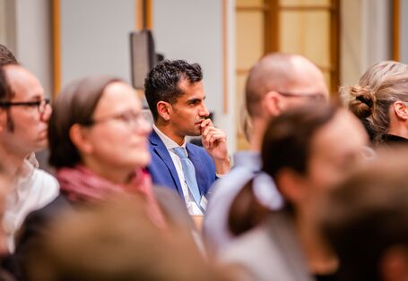 Der Blick geht durch die Teilnehmenden auf einen Teilnehmer, der sehr nachdenklich schaut. Er trägt einen blauen Anzug mit hellblauem Schlips und weisses Hemd. Der Raum ist hell und holzfarben abgesetzt.