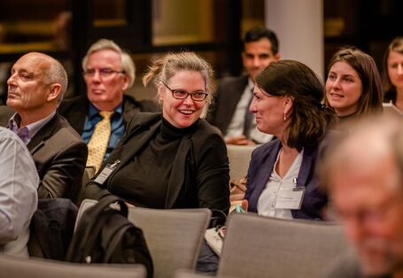 Teilnehmende sitzen im Veranstaltungsraum in Reihen und unterhalten sich lachend.