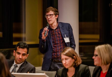 Teilnehmender steht im Veranstaltungsraum und spricht in ein Mikrofon, welches er in seiner rechten Hand hält.