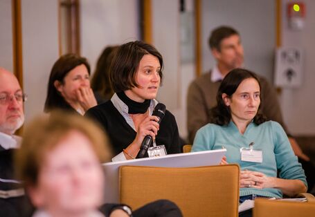 Teilnehmende sitzen im einem Veranstaltungsraum auf orangenen Stühlen in Reihen und blicken nachdenklich. Eine Teilnehmerin spricht in ein Handmikro.