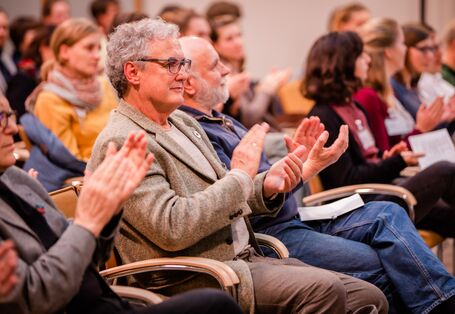 Teilnehmende sitzen auf orangenen Stühlen in Reihen im Veranstaltungsraum und applaudieren.