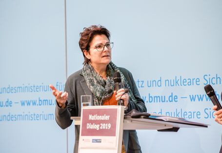 Zwei Vortragende stehen am Stehpult vor blauem Hintergrund, auf dem der Veranstaltungsort geschrieben steht. Beide halten Handmikrofone in den Händen und sprechen gestikulierend  miteinander.