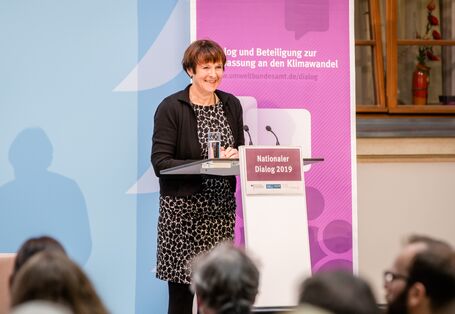 Teilnehmende hält eine Rede am Stehpult. Auf dem Stehpult steht ein Glas Wasser. Im Hintergrund steht ein Aufsteller sowie vorne auf dem Stehpult mit dem Veranstaltungstitel vorwiegend in lila gehalten.