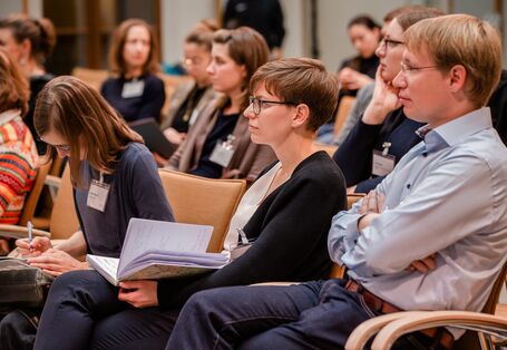 Teilnehmende sitzen im Veranstaltungsraum auf orangenen Stühle in Reihen und schauen zuhörend in eine Richtung.