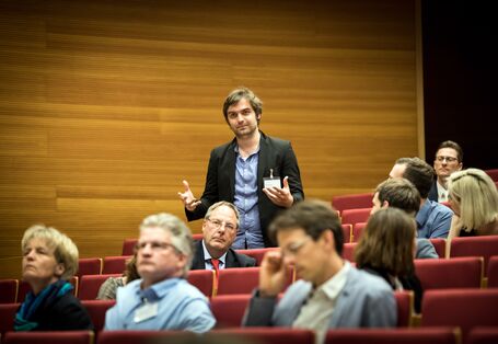  Teilnehmer steht präsent im Hörsaal innerhalb der roten Stuhlreihen und spricht. Die anderen Teilnehmerinnen und Teilnehmer sitzen rundherum und hören zu.