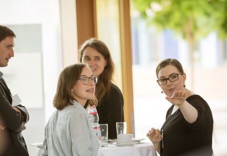 Teilnehmer und Teilnehmerinnen stehen in der Pause am Bistrotisch. Sie folgen schauend dem Hinweis einer Teilnehmerin, die in eine Richtung zeigt. Auf dem Bistrotisch stehen Gläser, Tassen und Teller sowie ein Getränkeflasche.Auf dem B