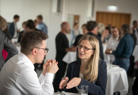 Teilnehmer und Teilnehmer stehen gemeinsam am Bistrotisch und unterhalten sich gestikulierend miteinander. Im Hintergrund sind stehen andere Teilnehmerinnen und Teilnehmer ebenfalls um den Bistrotischen herum.