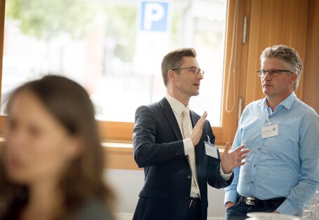 Zwei Teilnehmer stehen unterhaltend vor einem großen Fenster während der Pause am Bistrotisch.