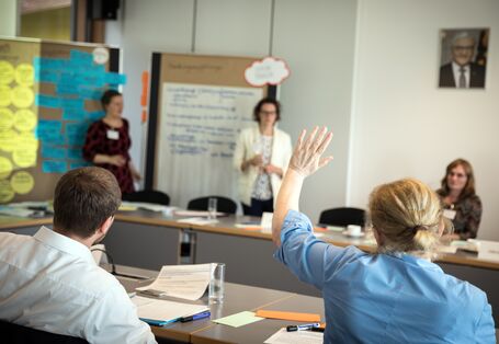 Zwei Teilnehmerinnen stehen vor zwei Moderationswänden. Die Wände sind mit Moderationsmaterialien bestückt. Im Vordergrund hebt eine Teilnehmerin in blauer Bluse die Hand zur Wortmeldung. Andere Teilnehmerinnen und Teilnehmer sitzen sich geben über am Arbeitstisch. Im Hintergrund hängt ein Bild des deutschen Bundespräsidenten.