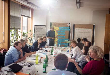 Teilnehmerinnen und Teilnehmer sitzen sich am Arbeitstisch gegenüber. Auf dem Tisch liegen Moderationsmaterialien, Unterlagen. Es stehen verteilt Getränke und ein Laptop. Im Hintergrund steht ein Teilnehmer vor den Moderationswänden. Die sitzenden Teilnehmerinnen und Teilnehmer diskutieren miteinander.
