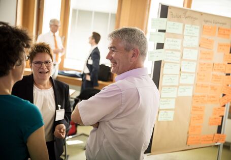 Drei Teilnehmerinnen und Teilnehmer stehen zusammen und schauen sich freundlich an und sprechen miteinander. Seitlich befindet sich eine mit Moderationskarten bestückte Moderationswand. Im Hintergrund sieht man ebenfalls Teilnehmer vor einem Fenster stehen, die sich miteinander sprechen.