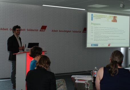 Vortragender steht am Rednerpult und präsentiert im Hintergrund das Programm des Stakeholderdialoges.