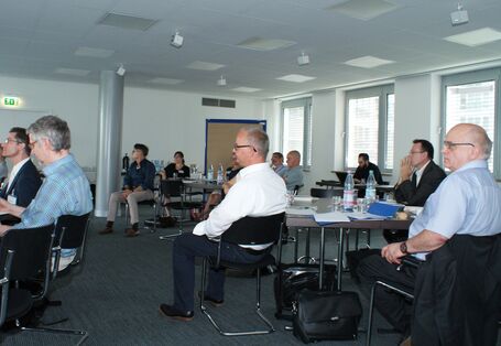 Teilnehmer sitzen im Arbeitsgruppenraum an Arbeitsgruppentischen mit dem Redner zugewandten Gesichtern.