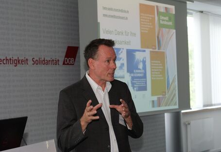 Vortragender steht am Rederpult und diskutiert gestikulierend mit den anderen Teilnehmern. Im Hintergrund ist der Abspann seiner Präsentation: Gesundheitliche Auswirkungen des Klimawandels in Deutschland