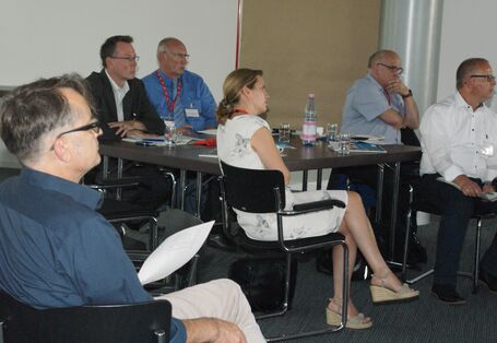 Teilnehmer sitzen im Arbeitsgruppenraum an Arbeitsgruppentischen mit dem Redner zugewandten Gesichtern.