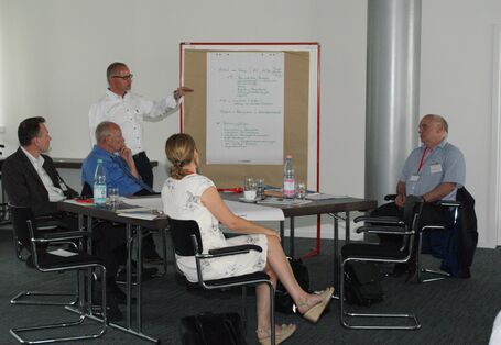 Vortragender steht an einer Moderationswand und erläutert die Arbeitsgruppenergebnisse, die dort handschriftlich angepinnt hängen.