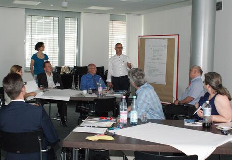 Vortragender steht an einer Moderationswand und trägt vor. Teilnehmer sitzen an den Gruppenarbeitstischen davor und hören aufmerksam zu.