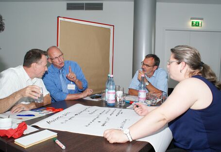 Teilnehmer diskutieren am Gruppenarbeitstisch. Eine Teilnehmerin schreibt Diskussionsergebnisse auf Posterpapier, welches auf dem Tisch liegt. Erfrischungsgetränke stehen auf dem Tisch. Ein Teilnehmer trinkt aus einem Glas Mineralwasser.