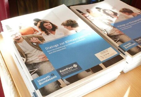 Das Bild zeigt zwei Stapel Broschüren mit dem Titel Dialoge zur Klimaanpassung. Eine Veranstaltung zur Chancen und Risiken des Klimawandels. Der Buchdeckel zeigt Menschen im Dialog. Eine Frau gibt dem Gesagten Ausdruck mit den Händen. Ein Mann nickt zustimmend im Hintergrund. Eine andere Frau hört zu.