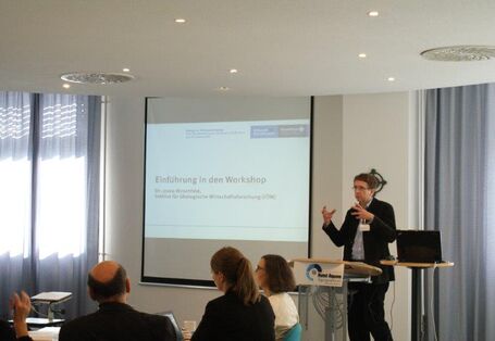 Ein Vortragender steht am Rednerpult und eröffnet gestikulierend den Dialog. Im Hintergrund erscheint das Eröffnungbild des Dialogs mit Titel und Logo.