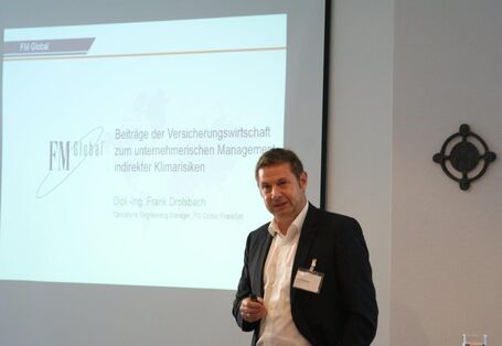 Vortragender steht mit dem Rücken vor seiner Präsentation im Hintergrund. Er hält einen Pointer in seiner Hand und spricht zu den Teilnehmern.