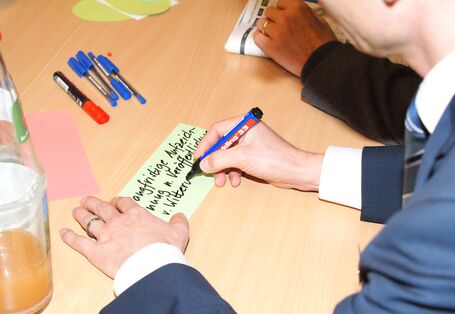 Teilnehmende beschriften farbige Moderationskarten in der Ansicht von oben sieht man die Hände. Farbige Moderationsstifte sowie eine Getränkeflasche sind ebenfalls zu sehen auf dem Gruppenarbeitstisch.