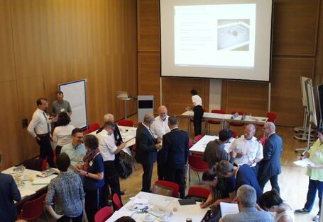 Teilnehmende stehen miteinander unterhaltend in lockeren Gesprächsgruppen beieinander in dem holzvertäfelten Veranstaltungsraum. Im Hintergrund stehen Rednerpult und die große Leinwand. Auf den Tischen befinden sich Arbeitsmaterialien und Getränke.