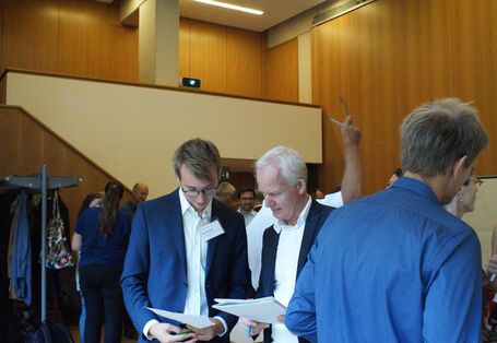 Zwei Teilnehmenden tauschen sich aus und betrachten dabei die Unterlagen in ihren Händen. Sie tragen blaue Sackos über weiße Hemden. 