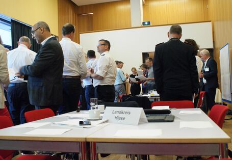 Teilnehemende stehen in lockeren Gesprächsgruppen beieinander. Sie tragen vorwiegend weiße Hemden und blaue Hosen. Im Vordergrund stehen ein Tischschild mit der Bezeichnung Landkreis Hiese.